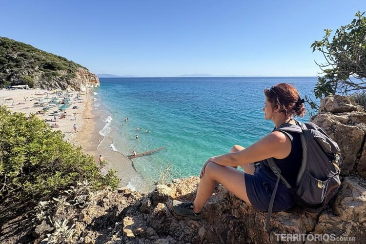 Mulher com mochila nas costas olha a paisagem do alto da montanha. Vista para a praia com banhistas, montanhas verdes e mar de cor azul como o céu.