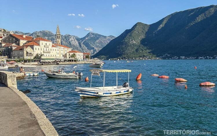 Barcos atracados em água azul da Baía de Kotor com cidade medieval e montanhas ao fundo. 
