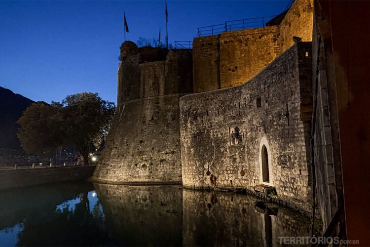 Muralha de cidade medieval iluminada reflete na água do lago, céu quase escuro.