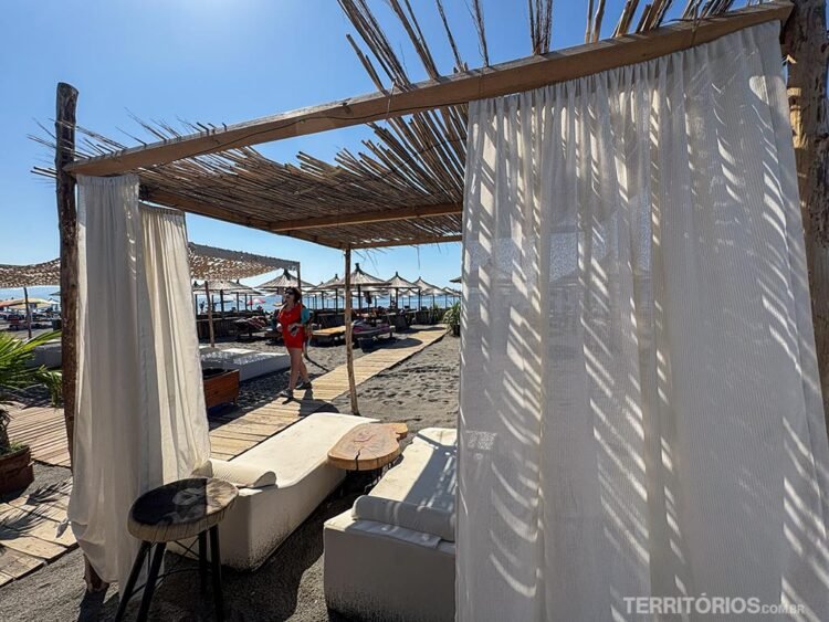Mulher veste roupa laranja e caminha em passarela de madeira para a praia. Espreguiçadeiras, sombreiros e cortinas no caminho. Céu azul