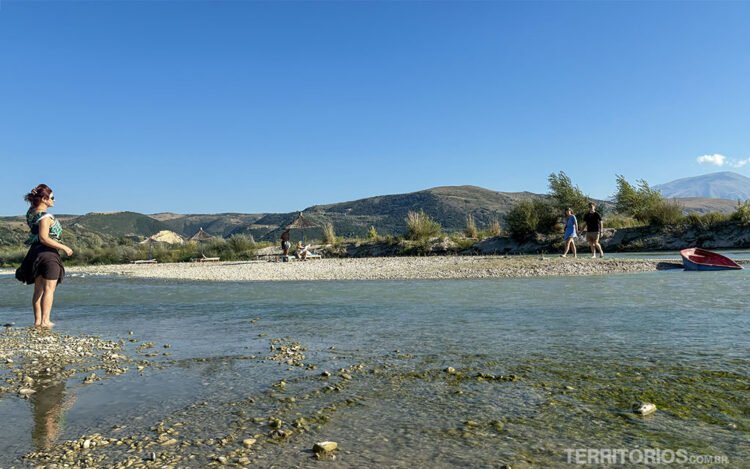Rio selvagem na Albânia entre montanhas e vegetação. Céu azul, um barco e pessoas caminham de pés descalços.