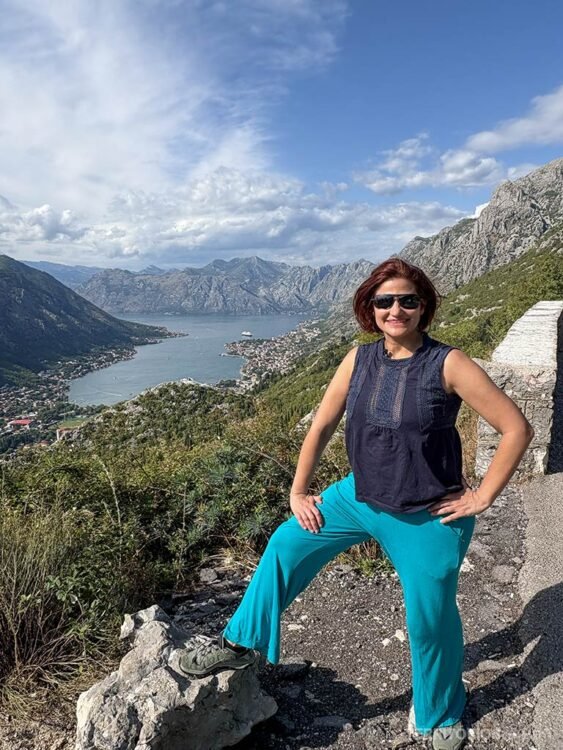 Mulher morena de óculos, blusa e calças azuis fazem pose em frente ao mirante para a Baía de Kotor 