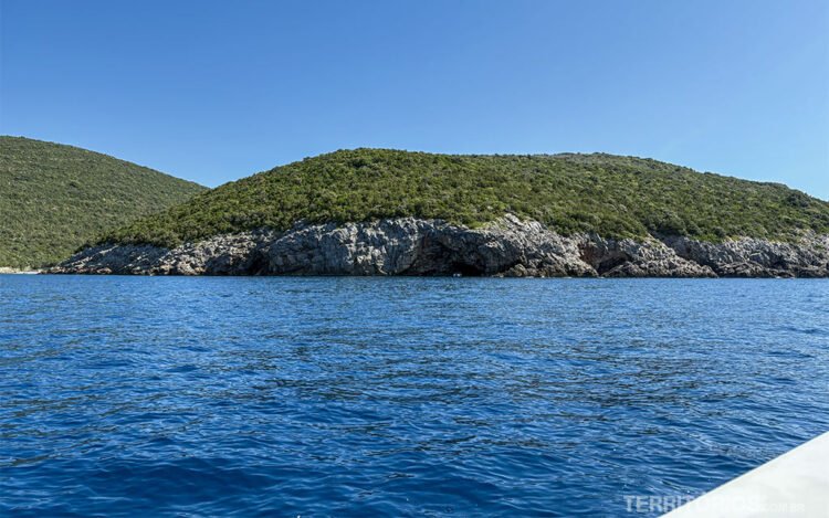 Baía de Kotor vista do barco. Montanha, caverna e vegetação em frente, céu azul.