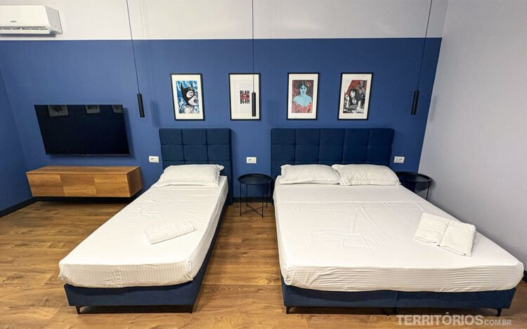 Quarto de hotel na capital da Albânia decorado com quadros na parede e tons de azul. Duas camas, uma de solteiro e outra de casal