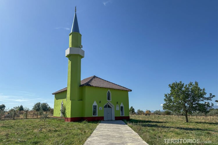 Mesquita de cor verde chamativa na zona rural da Albânia. Céu azul, gramado e árvores ao redor.