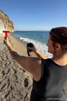 Mulher morena de maio preto segura letra T com uma mão e câmera com outra em cenário de praia com céu azul