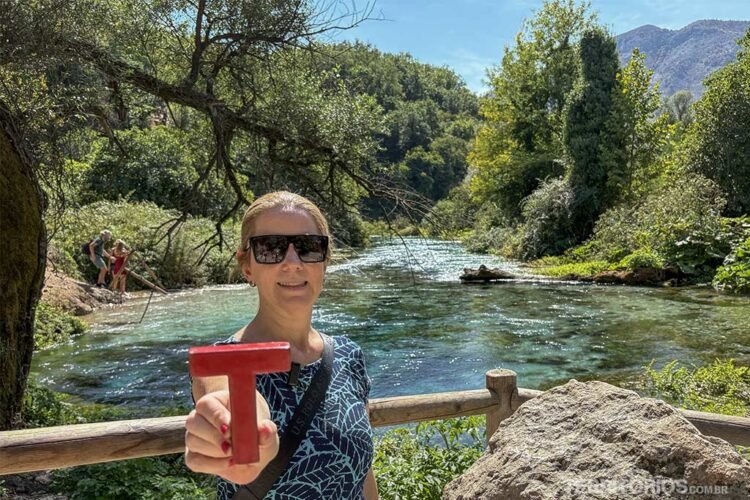 Mulher loira de óculos segura a letra T de madeira vermelha. Ao fundo lago verde e árvores