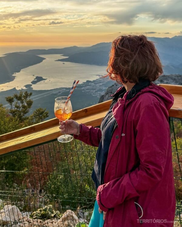 Mulher morena veste casaco magenta e olha o pôr do sol com drink na mão.